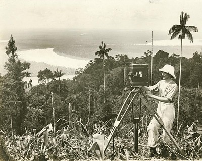 An old black-and-white photo of a man wearing a safari outfit. He stands behind a film camera that is placed on top of a sturdy tripod. In the background, you can see the jungle. The location is high up, as there is a horizon far behind the jungle.